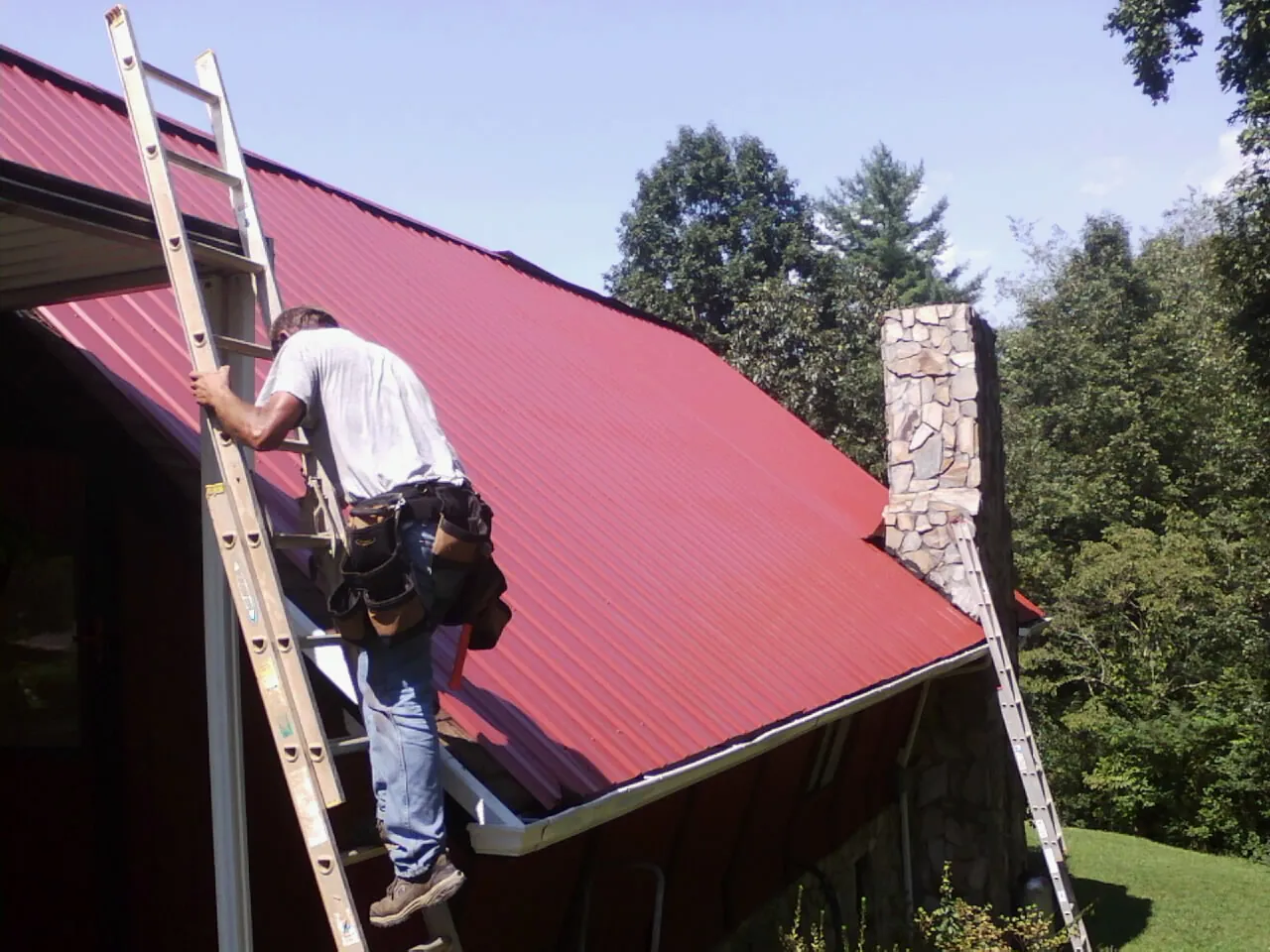 Roofer on a ladder beside a red metal roof and stone chimney — Soffit & Fascia Repair by Roofing Brothers in Hannibal
